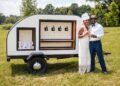 A joyful couple poses outdoors beside a stylish, white teardrop-shaped mobile bar trailer. The woman, on the left, wears a white tiered-fringe dress and a matching fascinator, while the man on the right is dressed in a white button-down shirt, a grey fedora, and blue trousers, holding a glass of champagne. The teardrop trailer features four beverage taps set against a white honeycomb backsplash and a framed "BAR" sign on the left. The scene is set on a sunny day in a grassy field with trees and a body of water in the background.