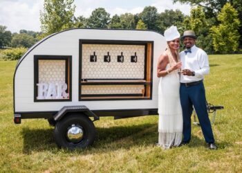 A joyful couple poses outdoors beside a stylish, white teardrop-shaped mobile bar trailer. The woman, on the left, wears a white tiered-fringe dress and a matching fascinator, while the man on the right is dressed in a white button-down shirt, a grey fedora, and blue trousers, holding a glass of champagne. The teardrop trailer features four beverage taps set against a white honeycomb backsplash and a framed "BAR" sign on the left. The scene is set on a sunny day in a grassy field with trees and a body of water in the background.