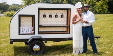 A joyful couple poses outdoors beside a stylish, white teardrop-shaped mobile bar trailer. The woman, on the left, wears a white tiered-fringe dress and a matching fascinator, while the man on the right is dressed in a white button-down shirt, a grey fedora, and blue trousers, holding a glass of champagne. The teardrop trailer features four beverage taps set against a white honeycomb backsplash and a framed "BAR" sign on the left. The scene is set on a sunny day in a grassy field with trees and a body of water in the background.