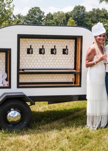 A joyful couple poses outdoors beside a stylish, white teardrop-shaped mobile bar trailer. The woman, on the left, wears a white tiered-fringe dress and a matching fascinator, while the man on the right is dressed in a white button-down shirt, a grey fedora, and blue trousers, holding a glass of champagne. The teardrop trailer features four beverage taps set against a white honeycomb backsplash and a framed "BAR" sign on the left. The scene is set on a sunny day in a grassy field with trees and a body of water in the background.