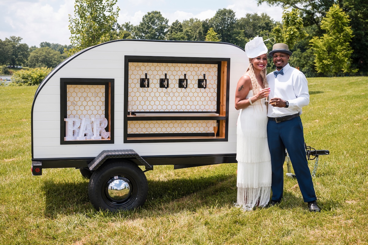 A joyful couple poses outdoors beside a stylish, white teardrop-shaped mobile bar trailer. The woman, on the left, wears a white tiered-fringe dress and a matching fascinator, while the man on the right is dressed in a white button-down shirt, a grey fedora, and blue trousers, holding a glass of champagne. The teardrop trailer features four beverage taps set against a white honeycomb backsplash and a framed "BAR" sign on the left. The scene is set on a sunny day in a grassy field with trees and a body of water in the background.