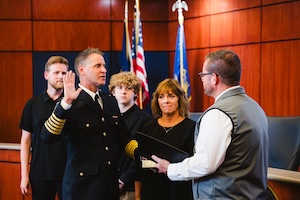 A medium shot captures the official swearing-in ceremony of Matt Mitchell as the Fire Chief of the Noblesville Fire Department. Chief Mitchell, dressed in a formal black dress uniform with gold stripes on his sleeves, stands on the left with his right hand raised, reciting the oath of office. He is surrounded by his family: his wife and two sons stand behind and beside him, watching with proud expressions. On the right, an official in a white shirt and gray vest holds a black folder while administering the oath. The ceremony takes place in a room with rich wood-paneled walls, with the American and Indiana state flags displayed in the background.