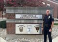 A medium full-length shot of a Noblesville police officer in a dark navy uniform standing next to the Noblesville Public Safety Building sign. The officer, identified as Chief Brad Arnold, is smiling at the camera with his hands at his sides, wearing a duty belt equipped with a radio, holster, and other tools. The sign is a large, rectangular structure made of red brick and topped with a gray stone ledge. Two metal plaques on the sign feature the words "NOBLESVILLE PUBLIC SAFETY BUILDING" and the logos for the Noblesville Police Department, City Court, and Noblesville Fire Department. The background shows the brick exterior of the building and trees with reddish-brown leaves under a clear sky.