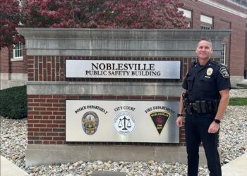 A medium full-length shot of a Noblesville police officer in a dark navy uniform standing next to the Noblesville Public Safety Building sign. The officer, identified as Chief Brad Arnold, is smiling at the camera with his hands at his sides, wearing a duty belt equipped with a radio, holster, and other tools. The sign is a large, rectangular structure made of red brick and topped with a gray stone ledge. Two metal plaques on the sign feature the words "NOBLESVILLE PUBLIC SAFETY BUILDING" and the logos for the Noblesville Police Department, City Court, and Noblesville Fire Department. The background shows the brick exterior of the building and trees with reddish-brown leaves under a clear sky.