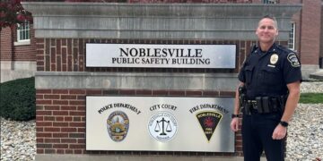 A medium full-length shot of a Noblesville police officer in a dark navy uniform standing next to the Noblesville Public Safety Building sign. The officer, identified as Chief Brad Arnold, is smiling at the camera with his hands at his sides, wearing a duty belt equipped with a radio, holster, and other tools. The sign is a large, rectangular structure made of red brick and topped with a gray stone ledge. Two metal plaques on the sign feature the words "NOBLESVILLE PUBLIC SAFETY BUILDING" and the logos for the Noblesville Police Department, City Court, and Noblesville Fire Department. The background shows the brick exterior of the building and trees with reddish-brown leaves under a clear sky.