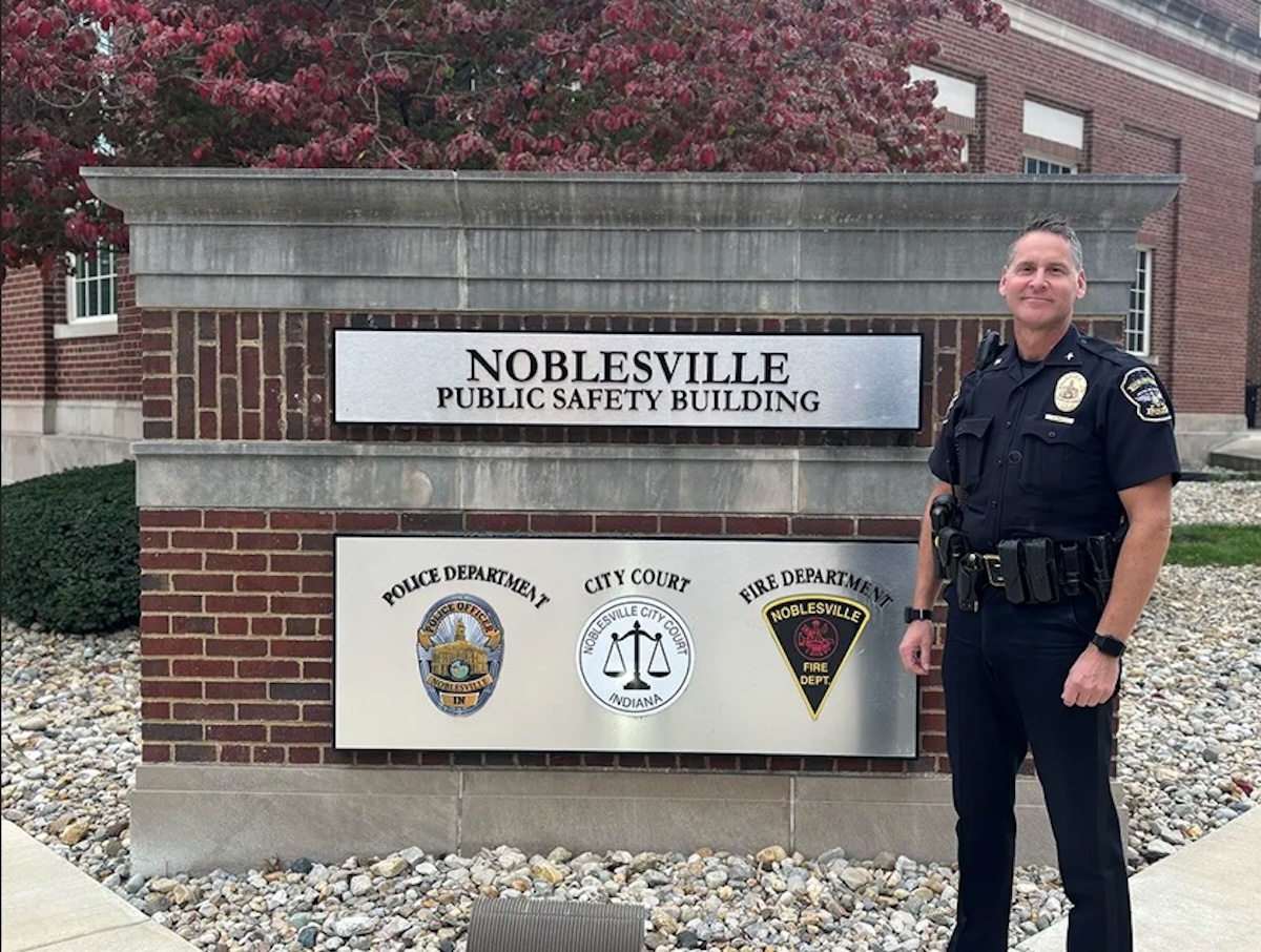 A medium full-length shot of a Noblesville police officer in a dark navy uniform standing next to the Noblesville Public Safety Building sign. The officer, identified as Chief Brad Arnold, is smiling at the camera with his hands at his sides, wearing a duty belt equipped with a radio, holster, and other tools. The sign is a large, rectangular structure made of red brick and topped with a gray stone ledge. Two metal plaques on the sign feature the words "NOBLESVILLE PUBLIC SAFETY BUILDING" and the logos for the Noblesville Police Department, City Court, and Noblesville Fire Department. The background shows the brick exterior of the building and trees with reddish-brown leaves under a clear sky.