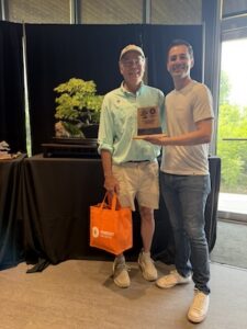 Two men stand smiling at an indoor bonsai exhibition. The man on the left, wearing a light blue polo shirt, a white baseball cap, and khaki shorts, holds a bright orange Pabst Blue Ribbon tote bag. The man on the right, in a light gray t-shirt and blue jeans, holds up a wooden award plaque for the camera. Behind them, a beautifully manicured bonsai tree with vibrant green foliage sits on a dark wooden display stand against a black backdrop.
