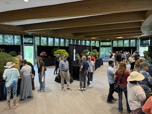 A wide, bright interior view of the Graeser Family Education Center at Waterfront Botanical Gardens in Louisville, KY. The modern, sustainable structure features high ceilings with prominent, parallel wooden glulam beams and floor-to-ceiling glass windows that look out onto lush greenery. The room is filled with attendees browsing a bonsai exhibition, with various miniature trees displayed on long, black-clothed tables. People of all ages are scattered throughout the open, wood-floored hall, creating a lively but serene atmosphere.