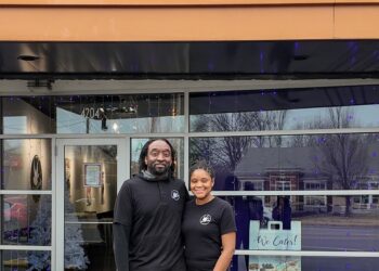 A man and a woman stand smiling together in front of a Cinnaholic Gourmet Cinnamon Rolls storefront. They are both dressed in black shirts featuring the company logo. The store has large glass windows reflecting the outdoors, and a "We Cater!" sign is visible in the window to their right. Above them, the large "CINNAHOLIC" sign is mounted on a dark, slatted exterior wall.