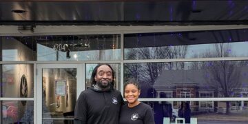 A man and a woman stand smiling together in front of a Cinnaholic Gourmet Cinnamon Rolls storefront. They are both dressed in black shirts featuring the company logo. The store has large glass windows reflecting the outdoors, and a "We Cater!" sign is visible in the window to their right. Above them, the large "CINNAHOLIC" sign is mounted on a dark, slatted exterior wall.