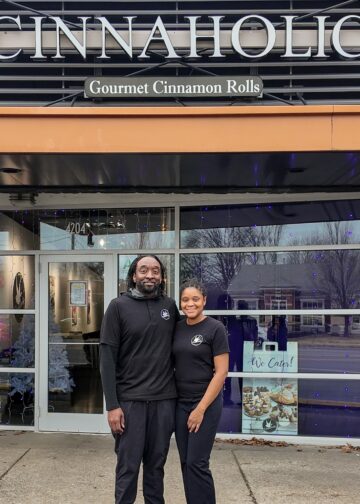 A man and a woman stand smiling together in front of a Cinnaholic Gourmet Cinnamon Rolls storefront. They are both dressed in black shirts featuring the company logo. The store has large glass windows reflecting the outdoors, and a "We Cater!" sign is visible in the window to their right. Above them, the large "CINNAHOLIC" sign is mounted on a dark, slatted exterior wall.
