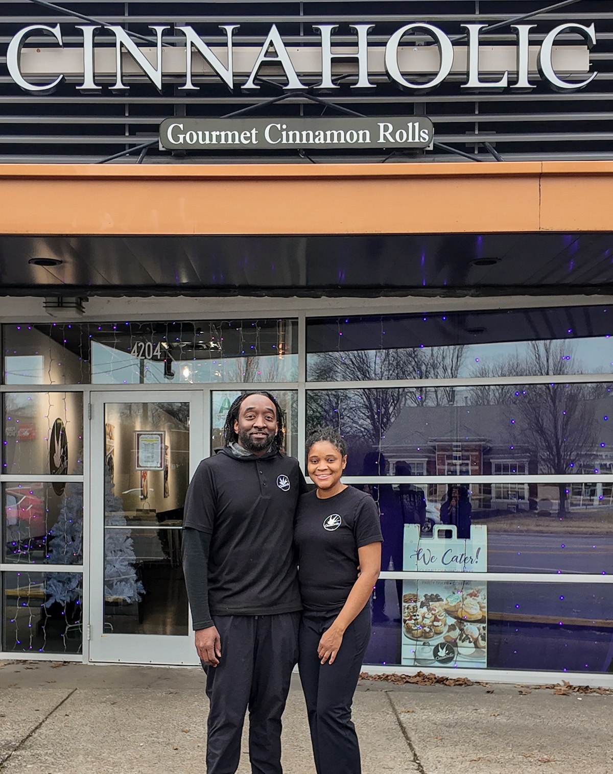 A man and a woman stand smiling together in front of a Cinnaholic Gourmet Cinnamon Rolls storefront. They are both dressed in black shirts featuring the company logo. The store has large glass windows reflecting the outdoors, and a "We Cater!" sign is visible in the window to their right. Above them, the large "CINNAHOLIC" sign is mounted on a dark, slatted exterior wall.