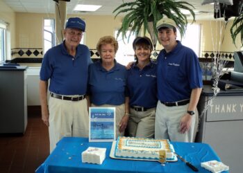 Four people, two men and two women of various ages, stand together behind a table in an ice cream shop or restaurant. They are all wearing matching blue polo shirts and khaki pants or shorts. On the table is a blue tablecloth, a decorated sheet cake, and a sign that reads "Founder's Day May 10, 1966." The background shows the interior of the shop with tiled walls and a large potted fern.