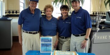 Four people, two men and two women of various ages, stand together behind a table in an ice cream shop or restaurant. They are all wearing matching blue polo shirts and khaki pants or shorts. On the table is a blue tablecloth, a decorated sheet cake, and a sign that reads "Founder's Day May 10, 1966." The background shows the interior of the shop with tiled walls and a large potted fern.