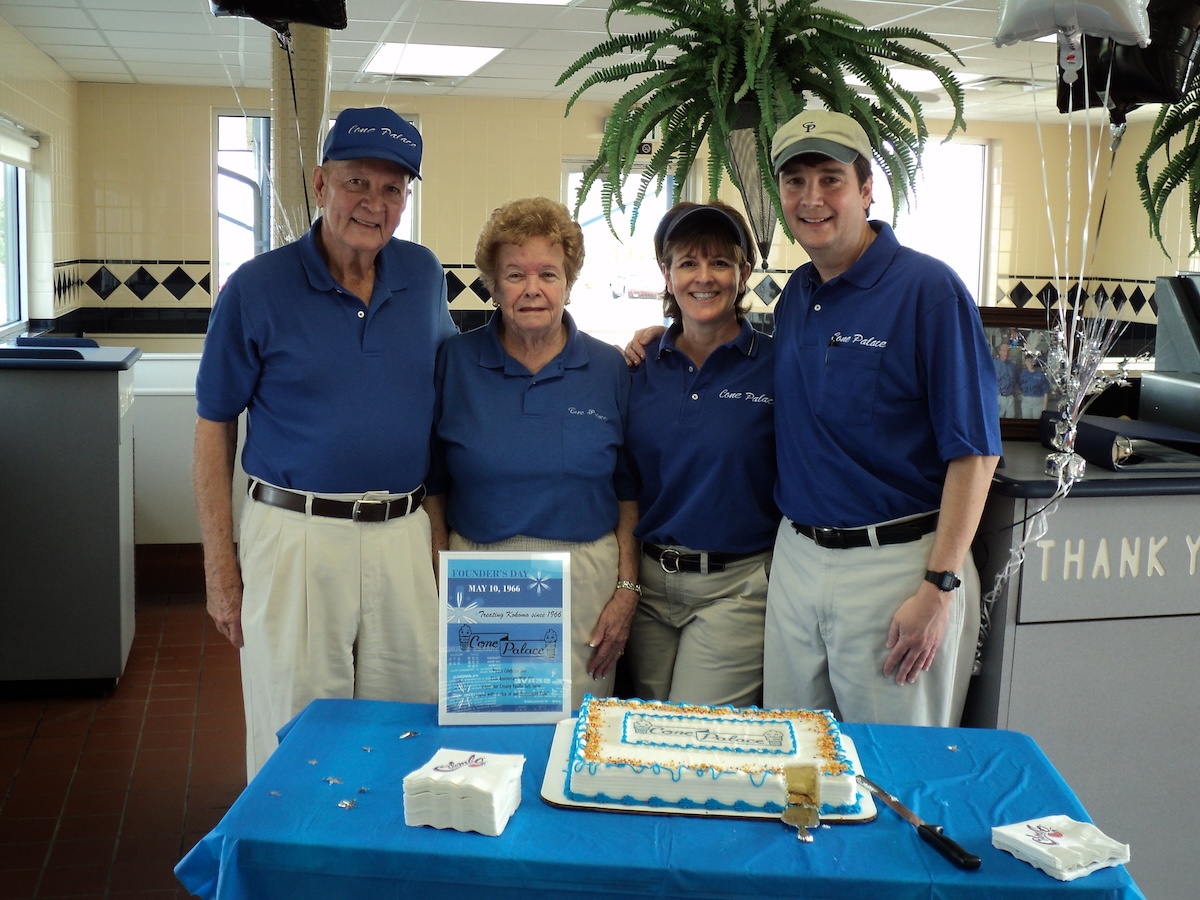 Four people, two men and two women of various ages, stand together behind a table in an ice cream shop or restaurant. They are all wearing matching blue polo shirts and khaki pants or shorts. On the table is a blue tablecloth, a decorated sheet cake, and a sign that reads "Founder's Day May 10, 1966." The background shows the interior of the shop with tiled walls and a large potted fern.