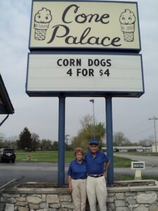 A low-angle vertical shot of an older man and woman standing together in front of a tall, blue-pillared sign for "Cone Palace." The woman is wearing a blue polo shirt and khaki pants, and the man wears a similar blue shirt, khaki pants, and a blue cap. The sign above them features two cartoon ice cream cones and a marquee that reads "CORN DOGS 4 FOR $4." The background shows a paved parking lot and a green field under an overcast sky.