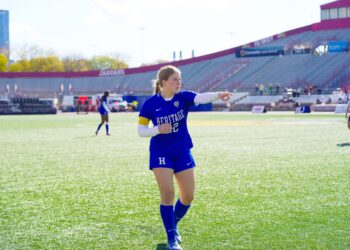A young woman with a long blonde ponytail, dressed in a royal blue Heritage Christian School soccer jersey with the number 32 and matching blue shorts, stands on a bright green turf field. She is wearing white compression sleeves on both arms and a captain's armband. She is looking and pointing toward the right side of the frame with her right arm extended. In the background, the red and gray grandstands of a large stadium are visible under a clear, sunny sky, with "JAGUARS" printed on one section of the seating.