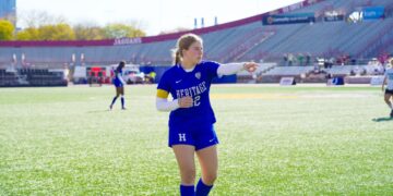 A young woman with a long blonde ponytail, dressed in a royal blue Heritage Christian School soccer jersey with the number 32 and matching blue shorts, stands on a bright green turf field. She is wearing white compression sleeves on both arms and a captain's armband. She is looking and pointing toward the right side of the frame with her right arm extended. In the background, the red and gray grandstands of a large stadium are visible under a clear, sunny sky, with "JAGUARS" printed on one section of the seating.