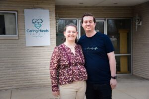 A man and a woman stand smiling together in front of the Caring Hands Clinic. The woman, on the left, is wearing a floral-patterned purple blouse and tan trousers. The man, on the right, is wearing a navy blue t-shirt with a white EKG heartbeat graphic. They are standing before a light-colored brick building, with the clinic's teal heart-and-hands logo visible on a sign behind them.