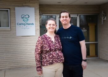 A man and a woman stand smiling together in front of the Caring Hands Clinic. The woman, on the left, is wearing a floral-patterned purple blouse and tan trousers. The man, on the right, is wearing a navy blue t-shirt with a white EKG heartbeat graphic. They are standing before a light-colored brick building, with the clinic's teal heart-and-hands logo visible on a sign behind them.