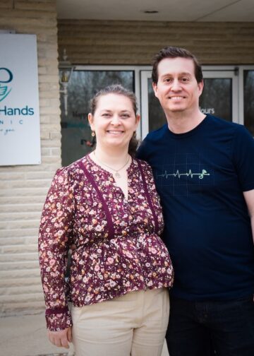A man and a woman stand smiling together in front of the Caring Hands Clinic. The woman, on the left, is wearing a floral-patterned purple blouse and tan trousers. The man, on the right, is wearing a navy blue t-shirt with a white EKG heartbeat graphic. They are standing before a light-colored brick building, with the clinic's teal heart-and-hands logo visible on a sign behind them.