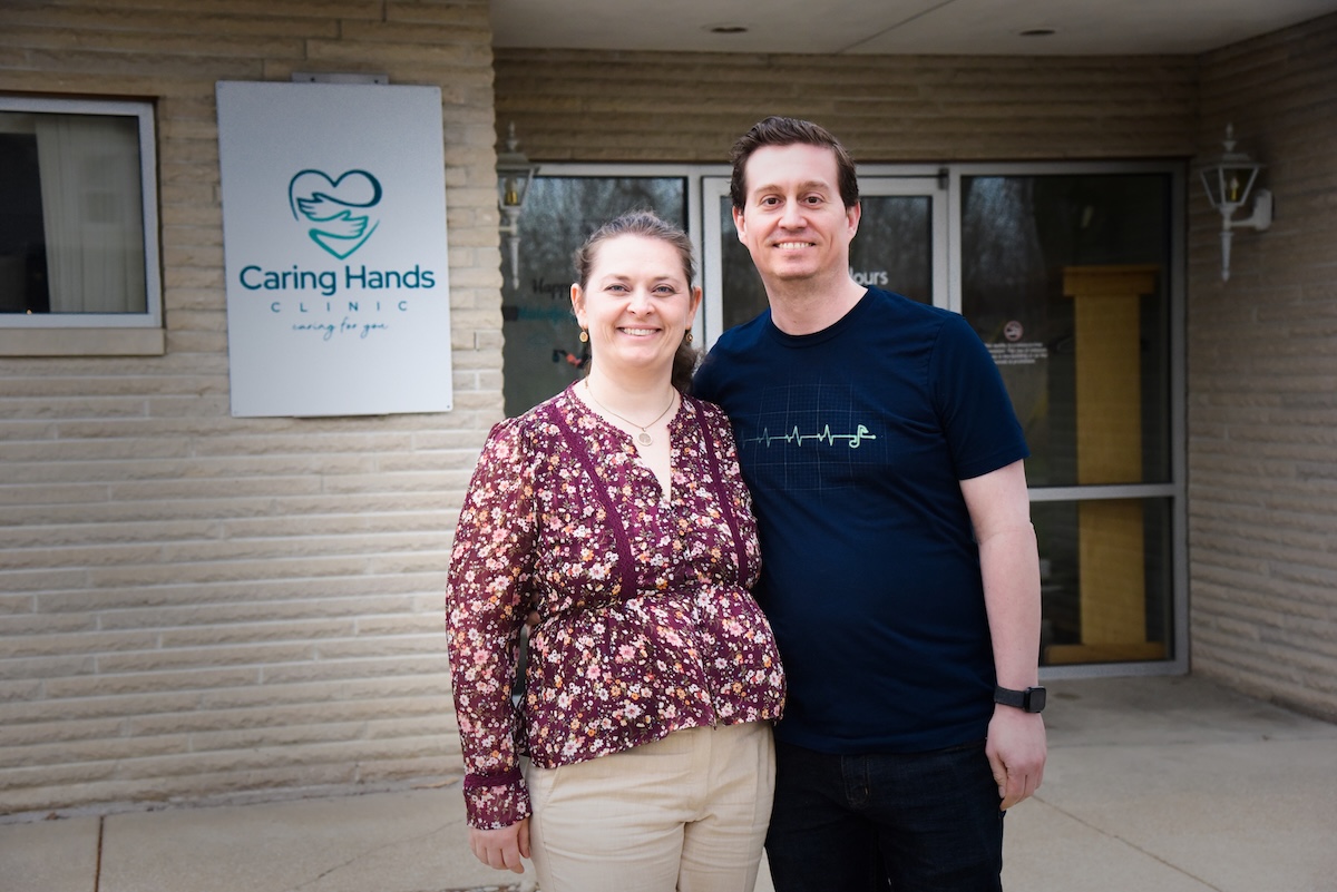 A man and a woman stand smiling together in front of the Caring Hands Clinic. The woman, on the left, is wearing a floral-patterned purple blouse and tan trousers. The man, on the right, is wearing a navy blue t-shirt with a white EKG heartbeat graphic. They are standing before a light-colored brick building, with the clinic's teal heart-and-hands logo visible on a sign behind them.