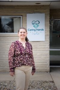 A professional portrait of a woman standing and smiling outdoors in front of a light-colored brick building. She is wearing a floral-patterned purple blouse and tan trousers. Directly behind her is a large white sign for "Caring Hands Clinic" featuring a teal heart-and-hands logo and the tagline "caring for you." The ground in front of the building is covered in small grey river stones.