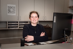 A woman with reddish-brown hair is sitting at an office desk, smiling at the camera with her arms crossed. She is wearing a dark, zip-up jacket. Her workspace includes a large Lenovo computer monitor, a desk organizer, and neutral-toned cabinetry in the background. A white paper with a large QR code is taped to the cabinet behind her.