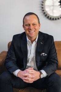 A man with short dark hair and a friendly smile, wearing a black blazer over a white dress shirt, sits on a brown leather couch with his hands clasped.