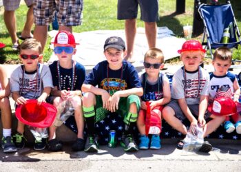 Seven young children sit in a row on a curb outdoors, many wearing patriotic American-themed clothing, red plastic fire hats, and sunglasses. Several children hold small bags of treats or plastic fire helmets, and one child in the center wears a black baseball cap and a navy blue "Survivor" t-shirt.