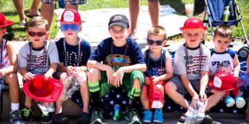 Seven young children sit in a row on a curb outdoors, many wearing patriotic American-themed clothing, red plastic fire hats, and sunglasses. Several children hold small bags of treats or plastic fire helmets, and one child in the center wears a black baseball cap and a navy blue "Survivor" t-shirt.