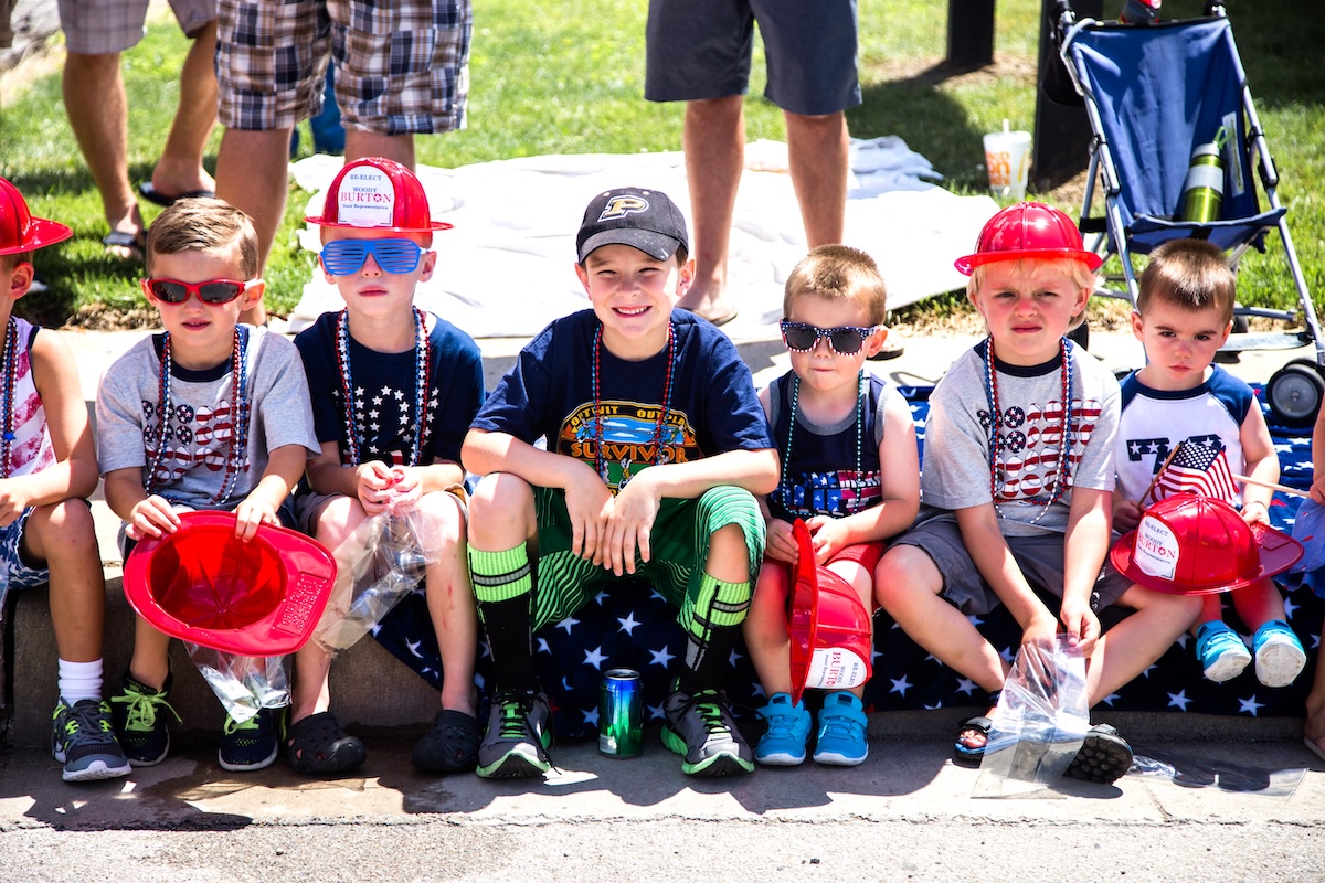 Seven young children sit in a row on a curb outdoors, many wearing patriotic American-themed clothing, red plastic fire hats, and sunglasses. Several children hold small bags of treats or plastic fire helmets, and one child in the center wears a black baseball cap and a navy blue "Survivor" t-shirt.