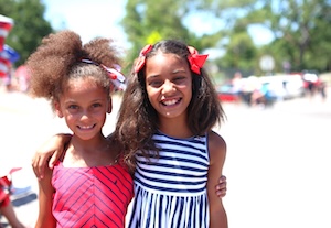 Two young girls smile for a photo outdoors. The girl on the left has her hair in a high puff with a patriotic-themed headband and wears a red patterned dress. The girl on the right has long dark hair with two large red bows and wears a navy blue and white striped sleeveless dress.