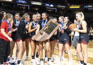 The Center Grove High School girls basketball team celebrates on the court after winning the 2026 IHSAA Class 4A state championship. A group of players in black jerseys and blue medals huddle together, laughing and cheering as two teammates in the center hold up a large, wooden state championship trophy. One player on the right holds a basketball, and the background shows the bright lights and seating of Gainbridge Fieldhouse.