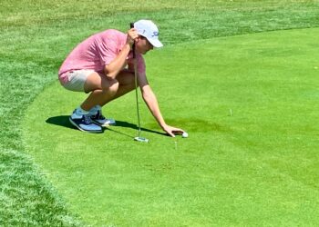 A young man in a pink patterned polo shirt, tan shorts, and a white Titleist baseball cap is crouching on a bright green golf green. He is carefully placing or adjusting a golf ball with his right hand while holding a putter in his left. Two white golf tees are visible on the grass nearby. The scene is set on a sunny day, with the sharp contrast of the neatly manicured green against a slightly taller fringe of grass.