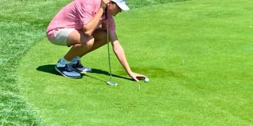 A young man in a pink patterned polo shirt, tan shorts, and a white Titleist baseball cap is crouching on a bright green golf green. He is carefully placing or adjusting a golf ball with his right hand while holding a putter in his left. Two white golf tees are visible on the grass nearby. The scene is set on a sunny day, with the sharp contrast of the neatly manicured green against a slightly taller fringe of grass.