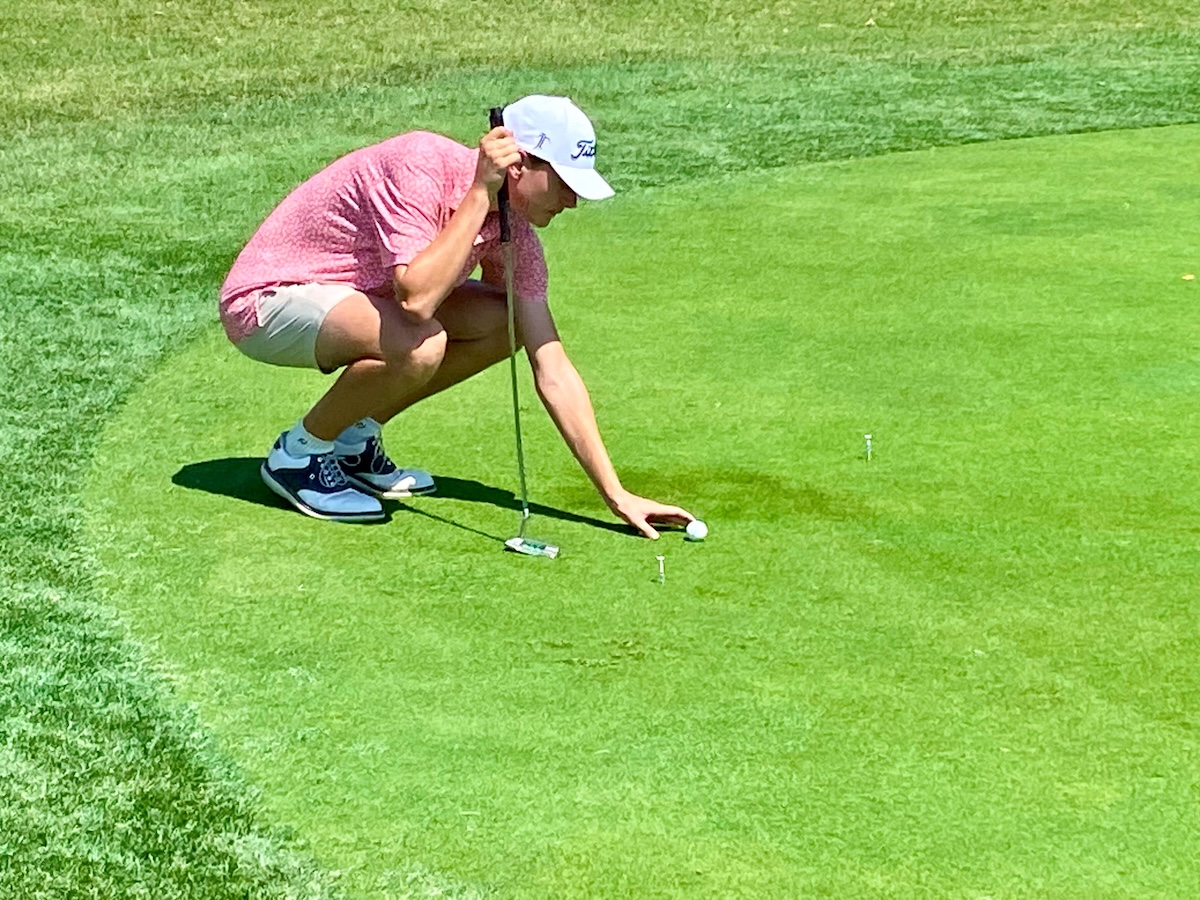 A young man in a pink patterned polo shirt, tan shorts, and a white Titleist baseball cap is crouching on a bright green golf green. He is carefully placing or adjusting a golf ball with his right hand while holding a putter in his left. Two white golf tees are visible on the grass nearby. The scene is set on a sunny day, with the sharp contrast of the neatly manicured green against a slightly taller fringe of grass.