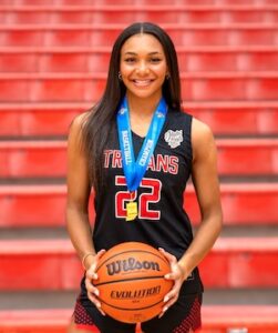 A smiling young female basketball player with long dark hair stands against a backdrop of red stadium seating. She is wearing a black Center Grove Trojans jersey with red and white lettering and the number "22." Around her neck, she wears a blue champion's medal, and she holds a Wilson Evolution basketball with both hands.