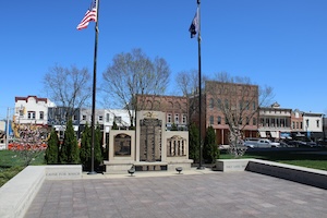 A wide-angle, eye-level shot of the Hamilton County Honor Roll, a war memorial located on the Courthouse Square in downtown Noblesville, Indiana. The memorial consists of three tiered stone tablets engraved with the names of local veterans, set within a paved plaza. Two tall flagpoles stand on either side: the left flying the American flag and the right flying a dark blue Indiana state flag. In the background, historic brick storefronts and a clear blue sky complete the serene downtown scene.