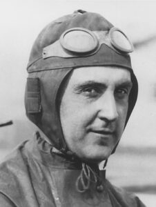 A black-and-white vintage portrait of Ray Harroun, the inaugural winner of the Indianapolis 500. He is wearing a dark, close-fitting leather aviator-style racing cap with a set of large, round goggles perched on his forehead. He has a slight, confident smile and is looking directly at the camera, dressed in a high-collared racing suit secured with a drawstring at the neck.