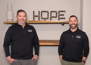 Two men wearing black quarter-zip pullovers with the Hope Plumbing logo stand in front of a neutral-colored wall. Behind them, a decorative "HOPE" sign made from industrial metal pipes sits on a wooden floating shelf.