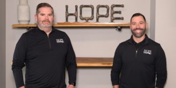Two men wearing black quarter-zip pullovers with the Hope Plumbing logo stand in front of a neutral-colored wall. Behind them, a decorative "HOPE" sign made from industrial metal pipes sits on a wooden floating shelf.