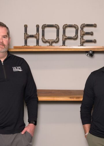 Two men wearing black quarter-zip pullovers with the Hope Plumbing logo stand in front of a neutral-colored wall. Behind them, a decorative "HOPE" sign made from industrial metal pipes sits on a wooden floating shelf.