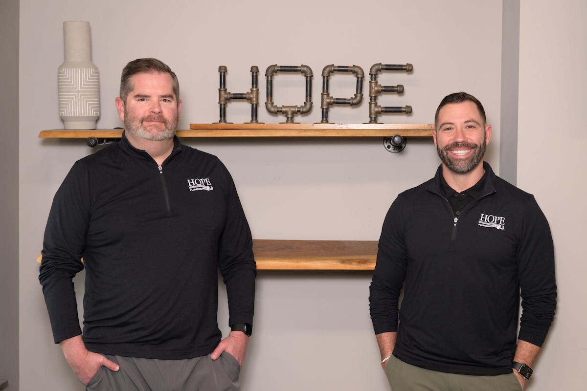 Two men wearing black quarter-zip pullovers with the Hope Plumbing logo stand in front of a neutral-colored wall. Behind them, a decorative "HOPE" sign made from industrial metal pipes sits on a wooden floating shelf.