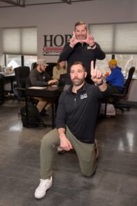 A lighthearted office photo featuring two men from Hope Plumbing in the foreground. One man is kneeling, pointing one finger upward, while the man standing behind him frames his face with his hands in a "camera" gesture. In the blurred background, other team members are working at desks in an open-concept office space under a large "HOPE" company logo.