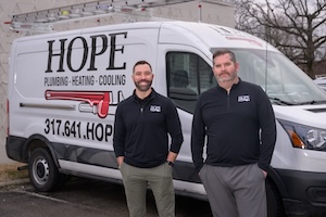 Two men in black logo pullovers stand confidently in front of a white Hope Plumbing, Heating, & Cooling service van. The van features a large "HOPE" logo and a red pipe wrench graphic above the company's contact number.