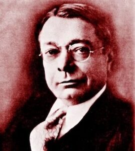 A sepia-toned vintage portrait of Ray Harroun, the legendary engineer and driver who won the inaugural Indianapolis 500 in 1911. He is wearing a formal dark suit, white collared shirt, and a wide silk tie. Harroun, nicknamed the "Little Professor" for his technical expertise, wears round, dark-rimmed glasses and looks directly at the camera with a steady, thoughtful expression.