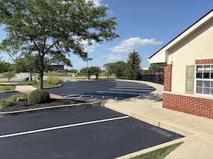 A wide view of a freshly sealed asphalt parking lot with crisp white parking lines, located in front of a tan and brick commercial building on a clear, sunny day.