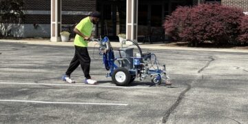 A man pushes a professional line-stripping machine across an asphalt parking lot in front of a brick building with a covered entrance; new white parking lines are visible on the ground.