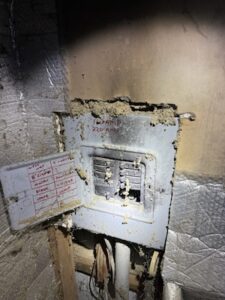 A dimly lit, close-up photo of an old, damaged electrical panel in a basement or crawlspace. The grey metal box is partially covered in clumps of yellow spray foam and debris. Its small door is open, revealing a handwritten circuit directory on the inside. Red lettering on the face of the panel reads "100 AMP 220 VOLT." The surrounding area shows exposed wooden studs and silver foil insulation, all appearing dusty and poorly maintained.